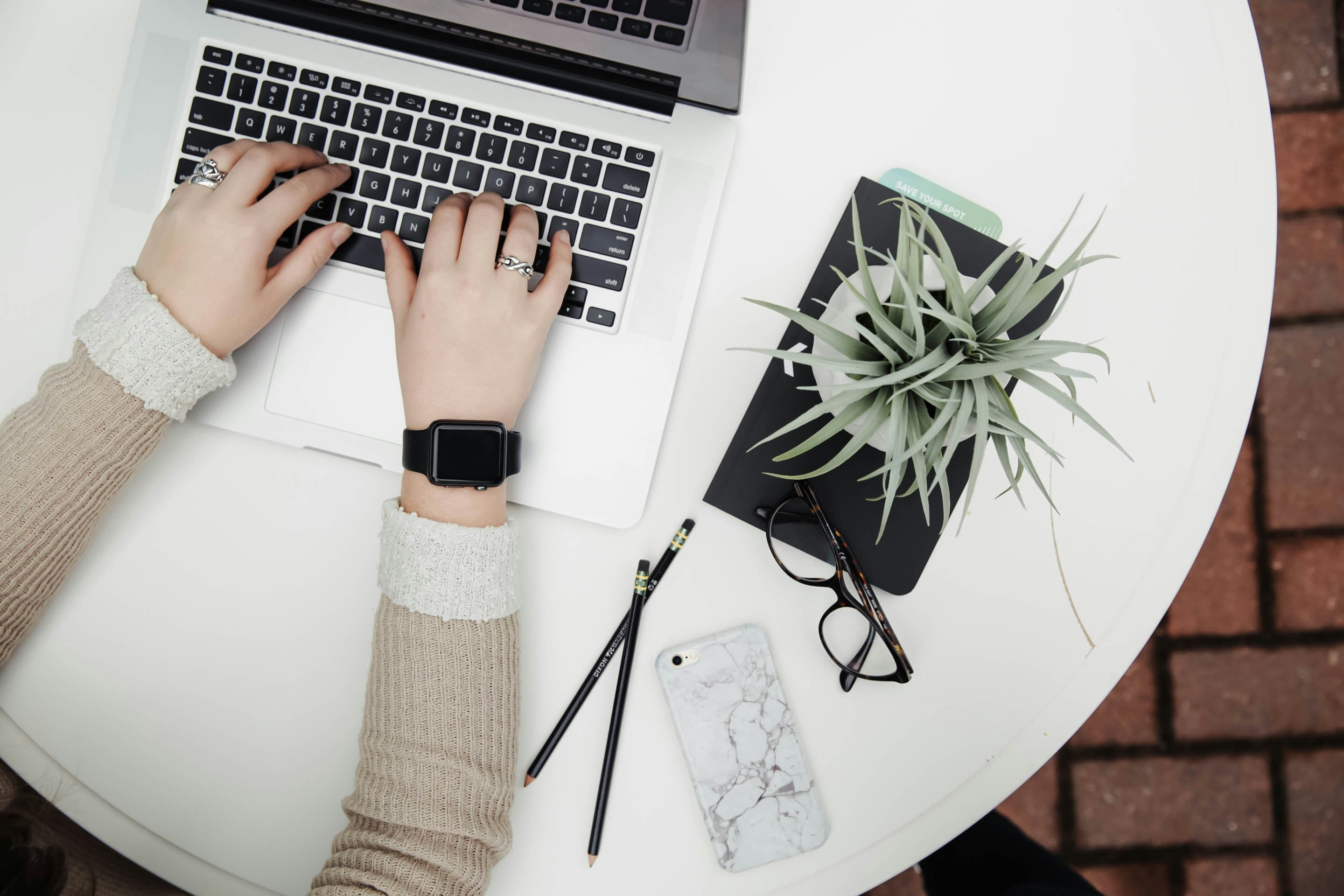 Hands typing on a laptop at a tidy desk with glasses, a plant and a phone